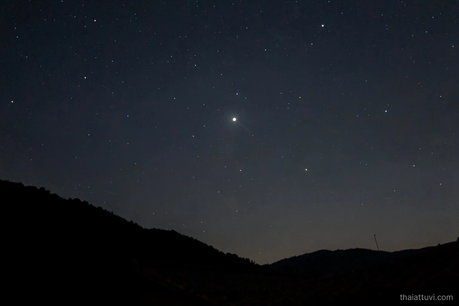 A night sky over a dark hillside with one very bright star, several fainter ones and a soft haze of tiny stars, silhouette of a tiled roof