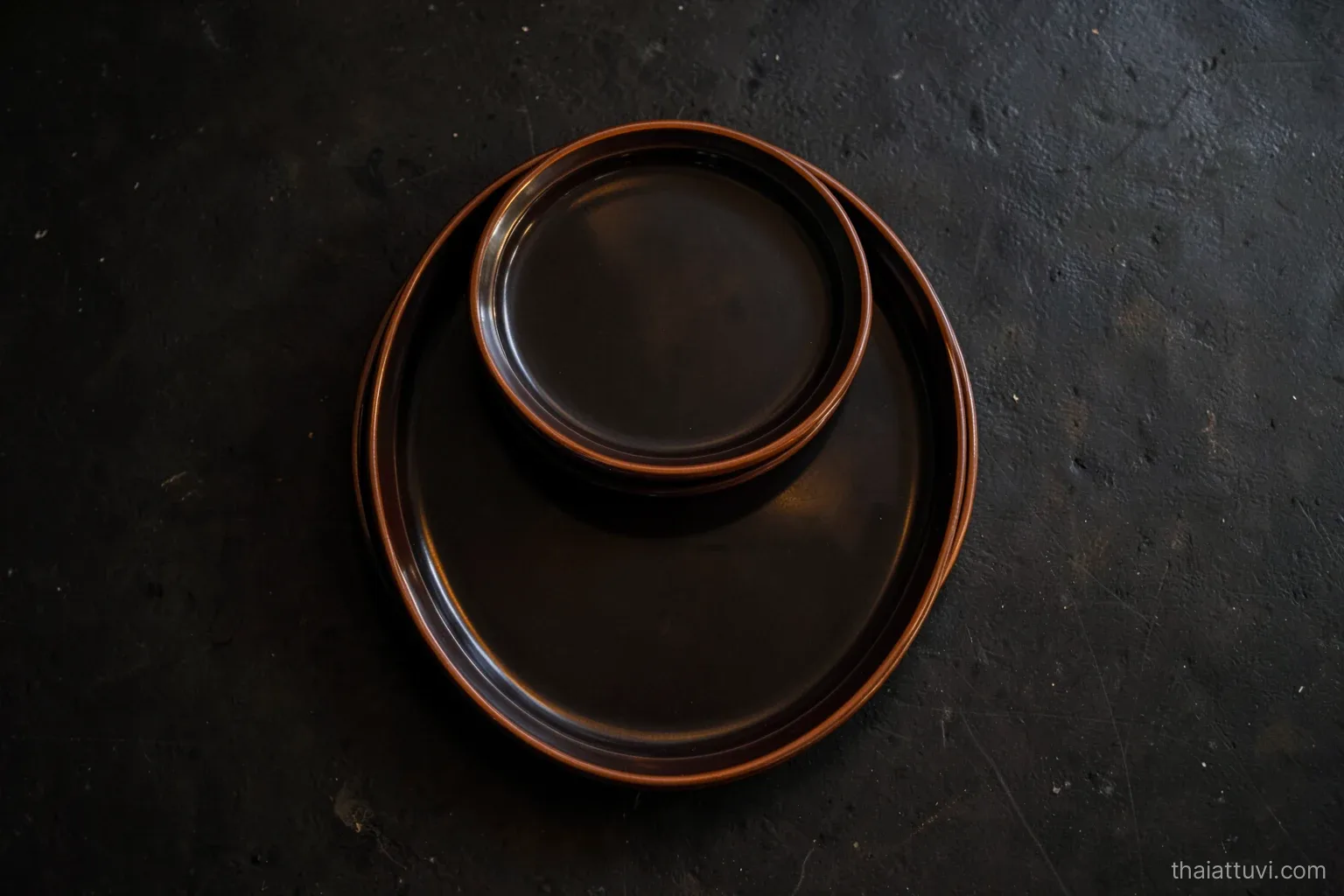 Three round lacquered trays of different sizes stacked on a dark floor, viewed from directly above, concentric rims glowing softly in warm lamplight